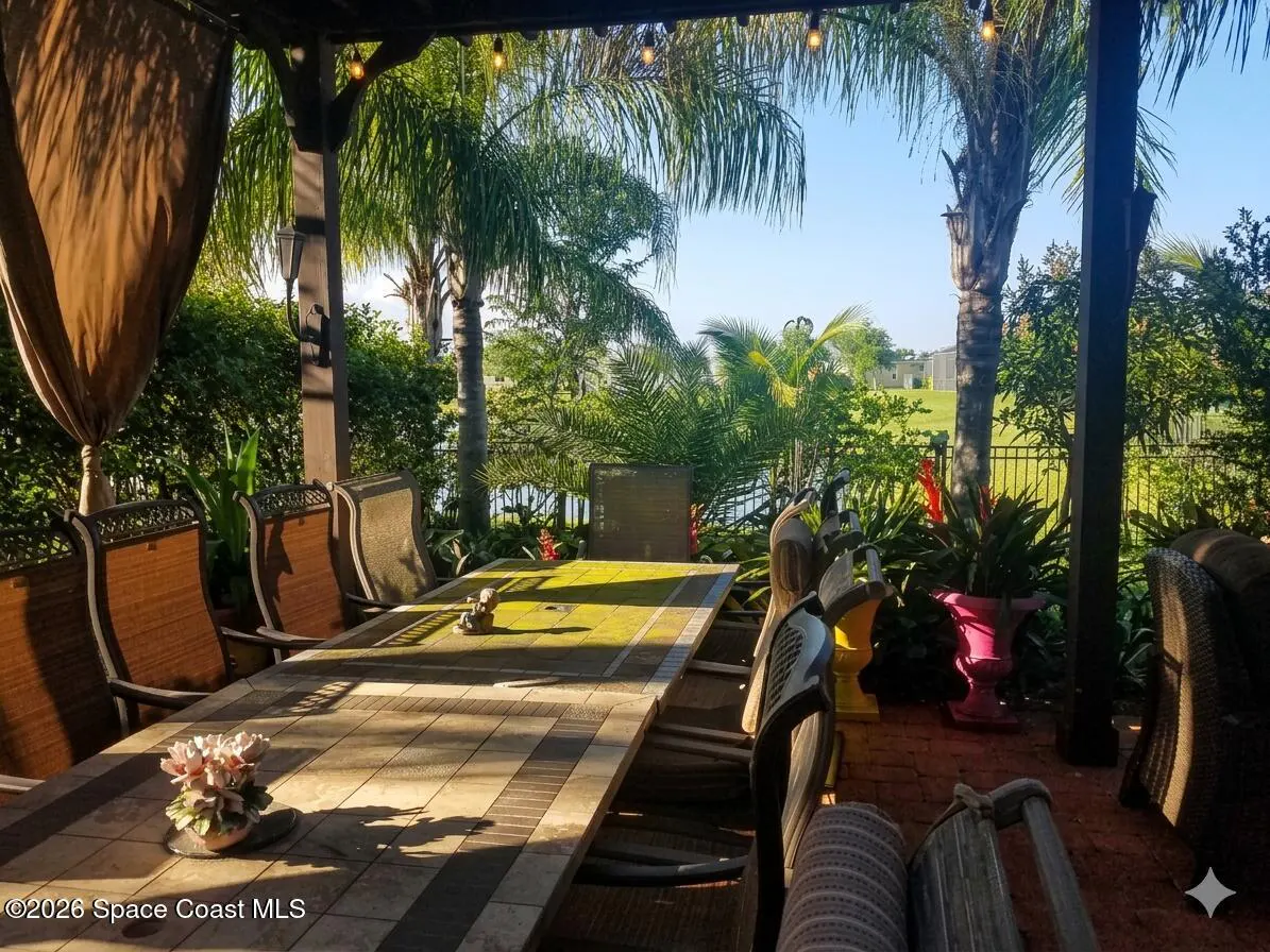 Outdoor patio with a long tile table and chairs under a pergola, overlooking a green lawn, palm trees, and a lake.