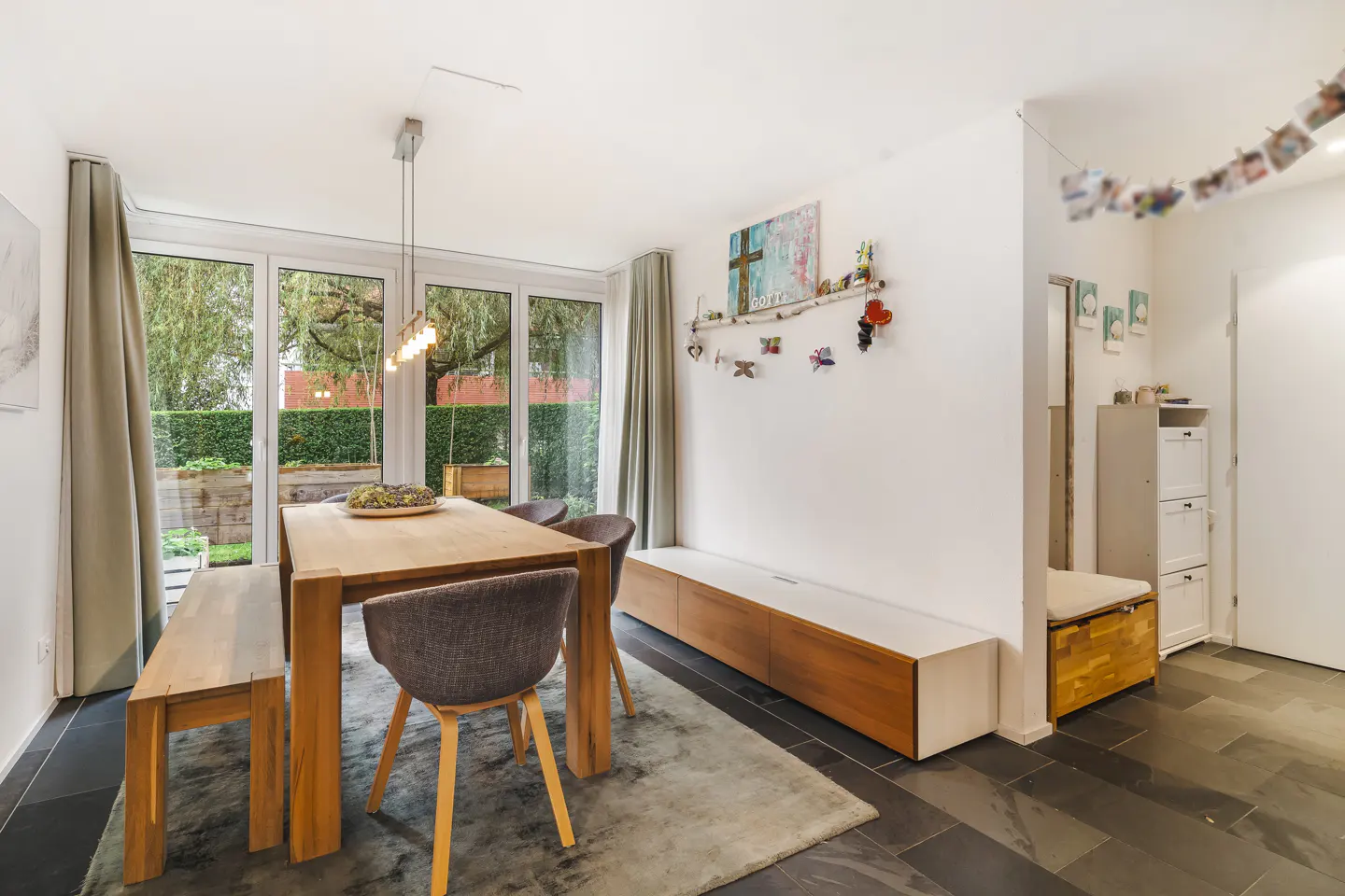 Bright dining room with a wooden table, grey chairs, and bench. Large windows overlook a green garden. Modern decor and slate flooring.