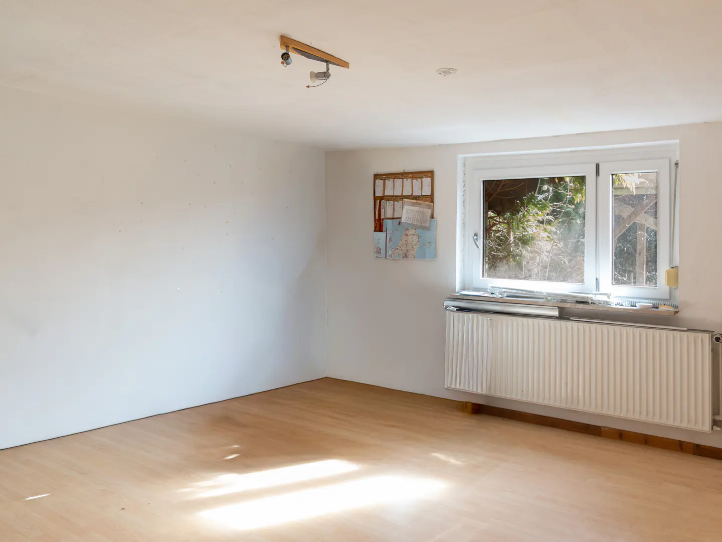 Bright, empty room with white walls, light wood floor, radiator under window, and corkboard on wall.