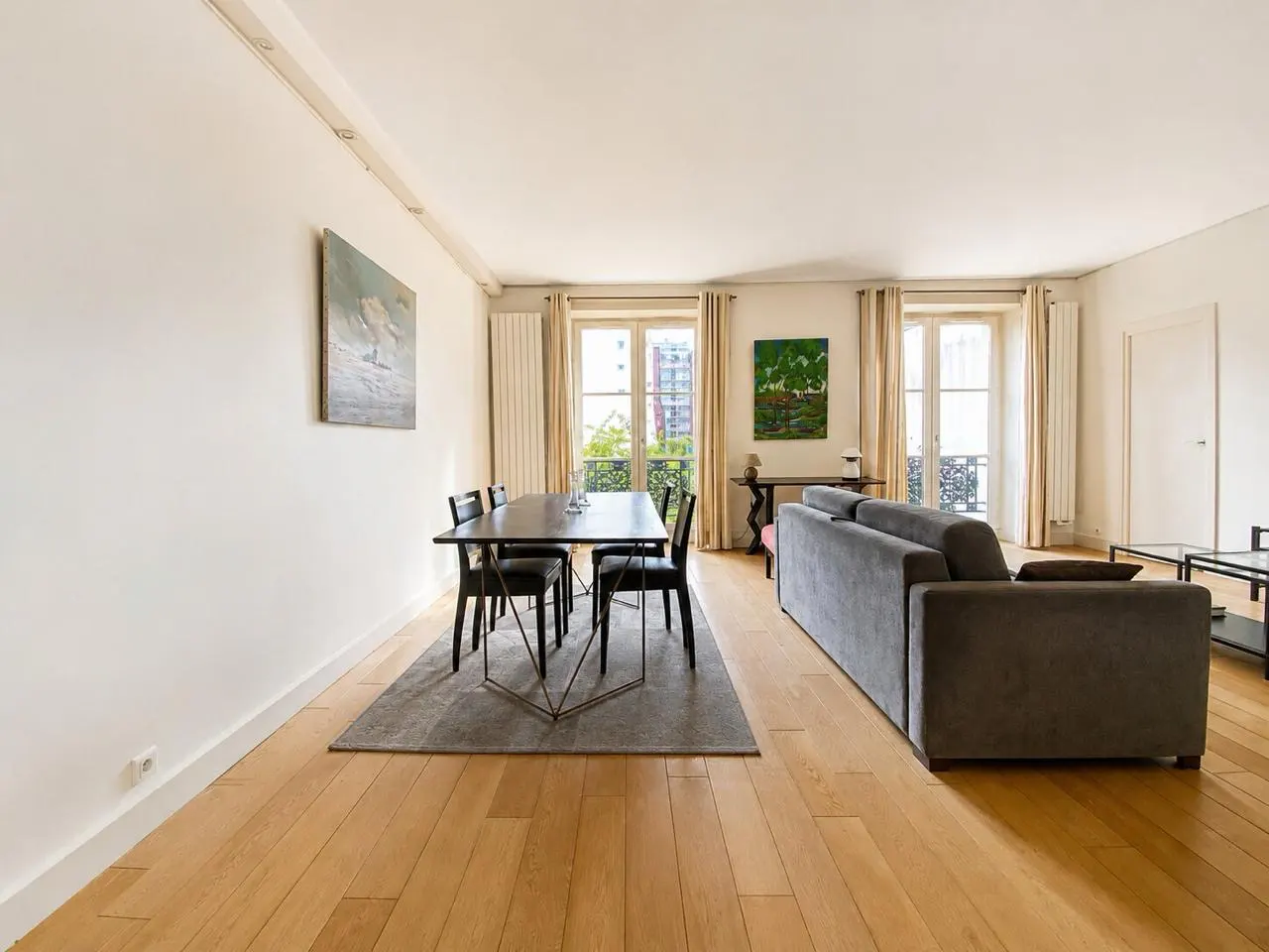 Bright living and dining room with hardwood floors, a grey sofa, and a dining table with four chairs near balcony doors.