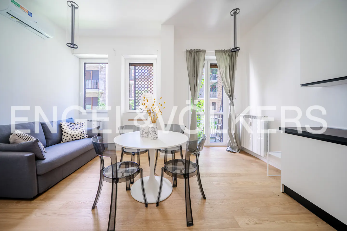 Modern living room with a grey sofa, white dining table, four transparent chairs, and light wood floors.