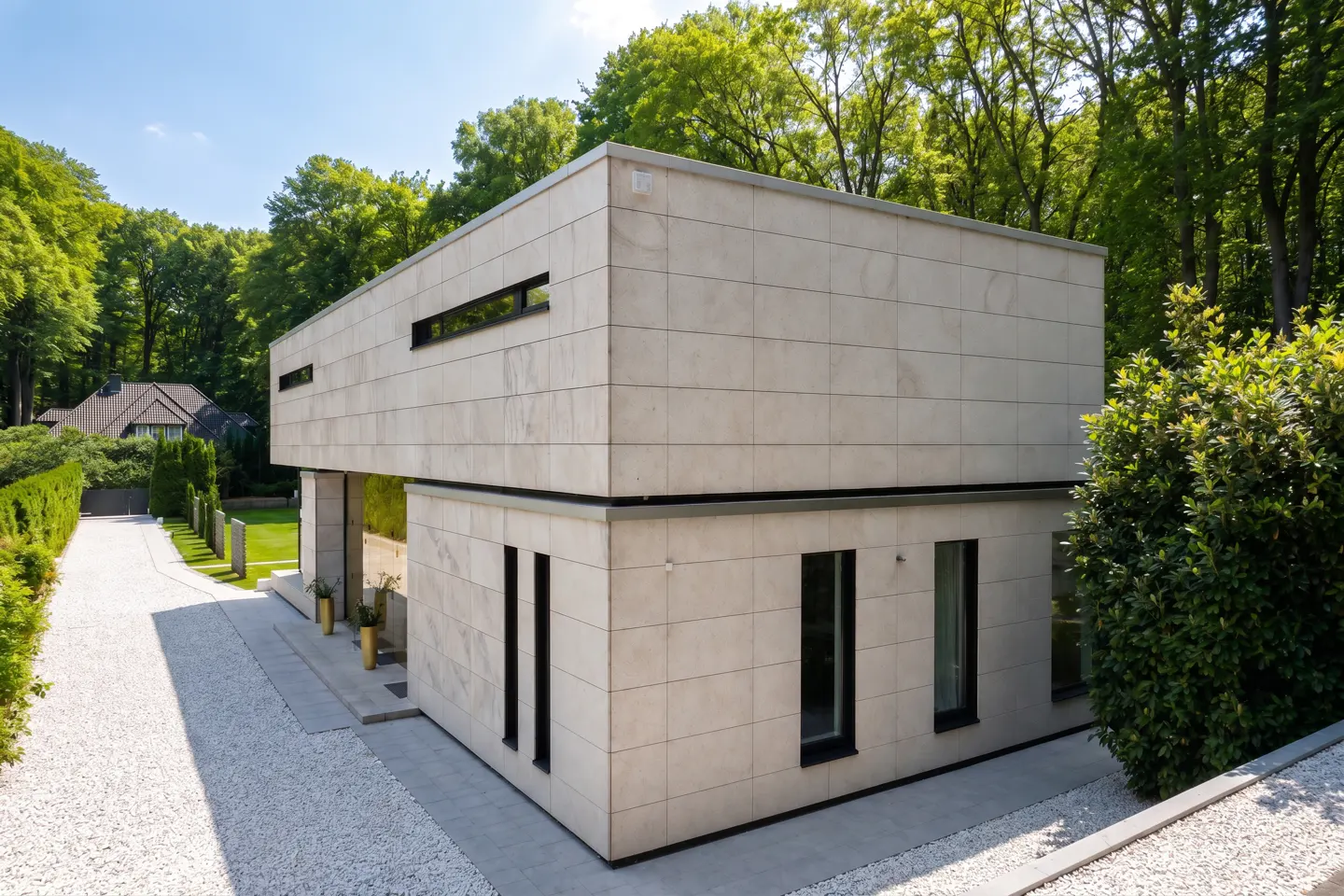 Two-story modern home with light stone exterior, black framed windows, and gravel driveway surrounded by green trees.