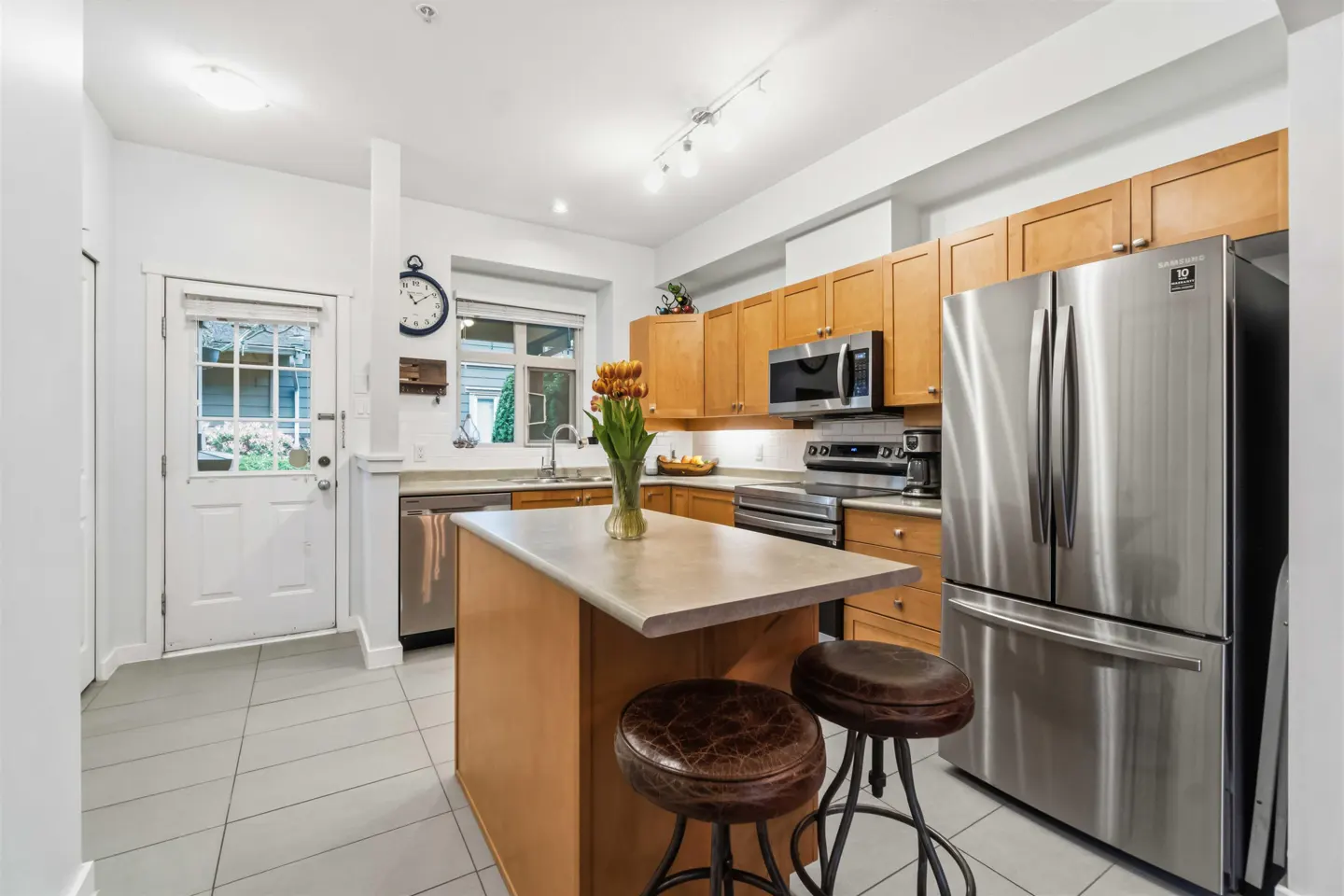 Bright kitchen with light wood cabinets, stainless steel appliances, and a center island with two stools. A vase of tulips sits on the island.