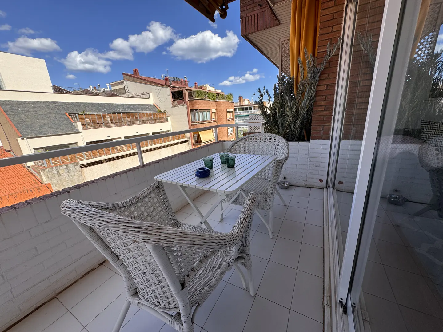 A balcony with white wicker furniture, including a table and chairs, overlooks a cityscape under a blue sky with clouds.