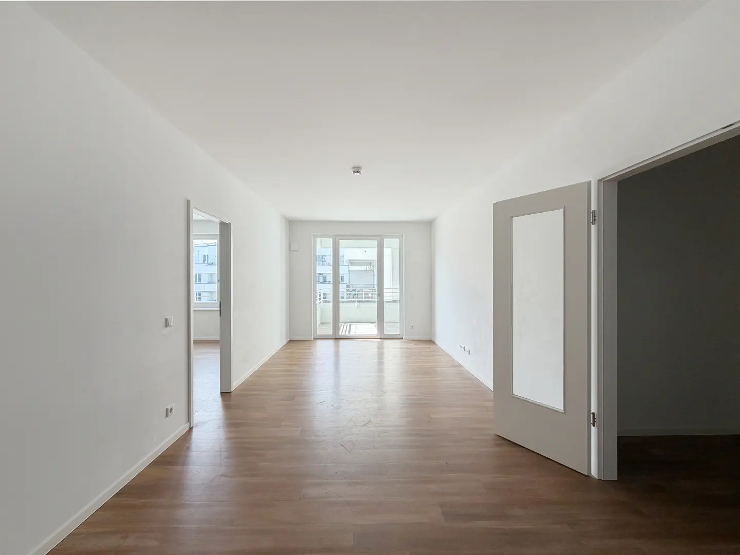 Bright, empty room with white walls, wood floors, and open doors. Balcony with white railings visible through glass doors at the far end.