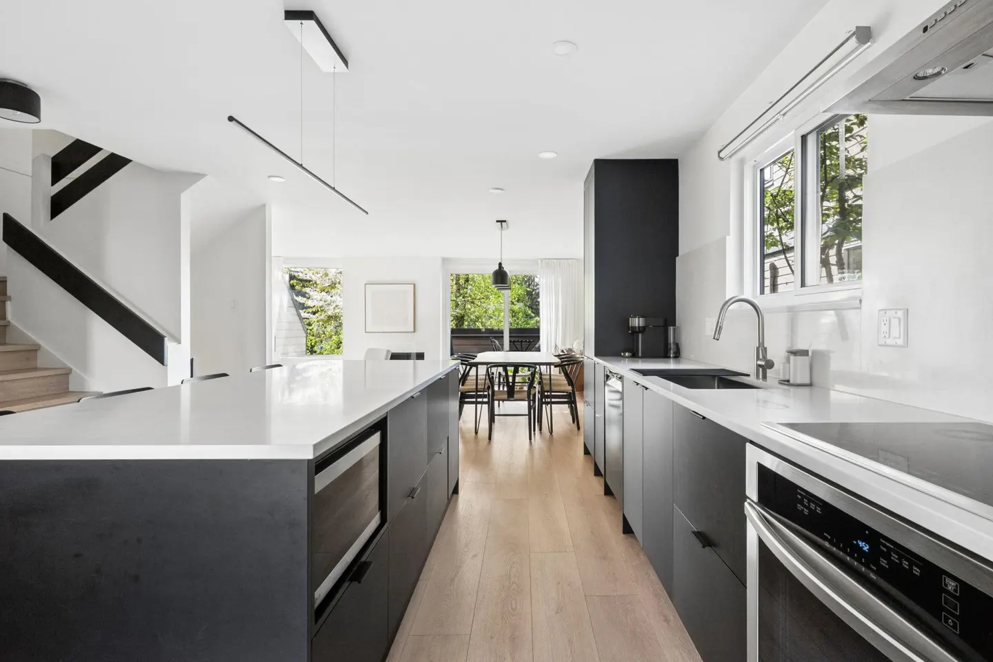 Modern kitchen with gray cabinets, white countertops, and light wood floors. A dining table is visible through a doorway.