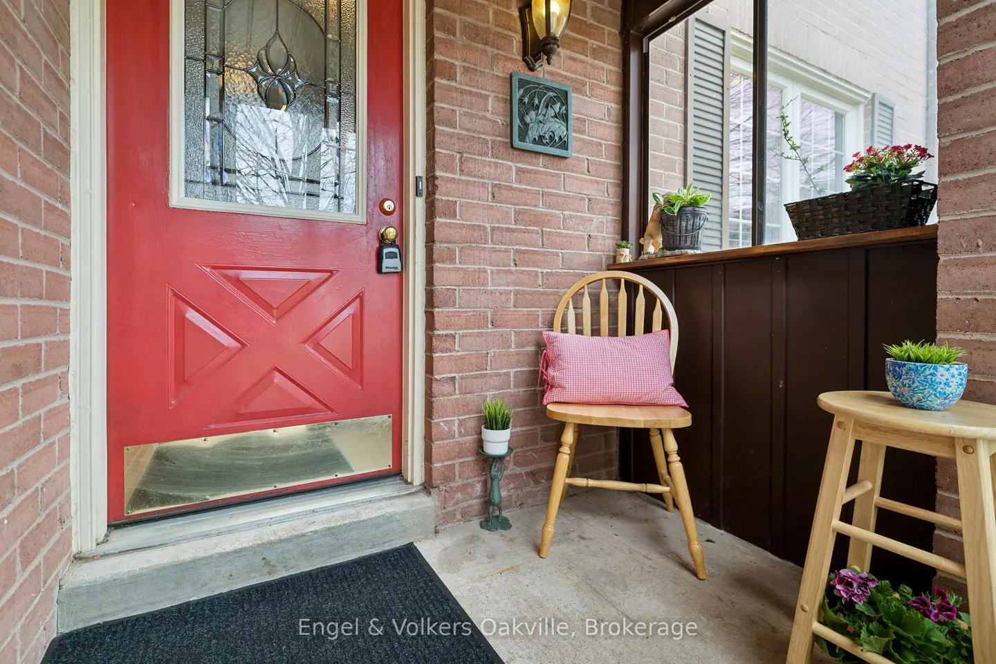 A red front door with a glass window, a wooden chair with a pink pillow, and a small wooden table with a blue pot on a brick porch.