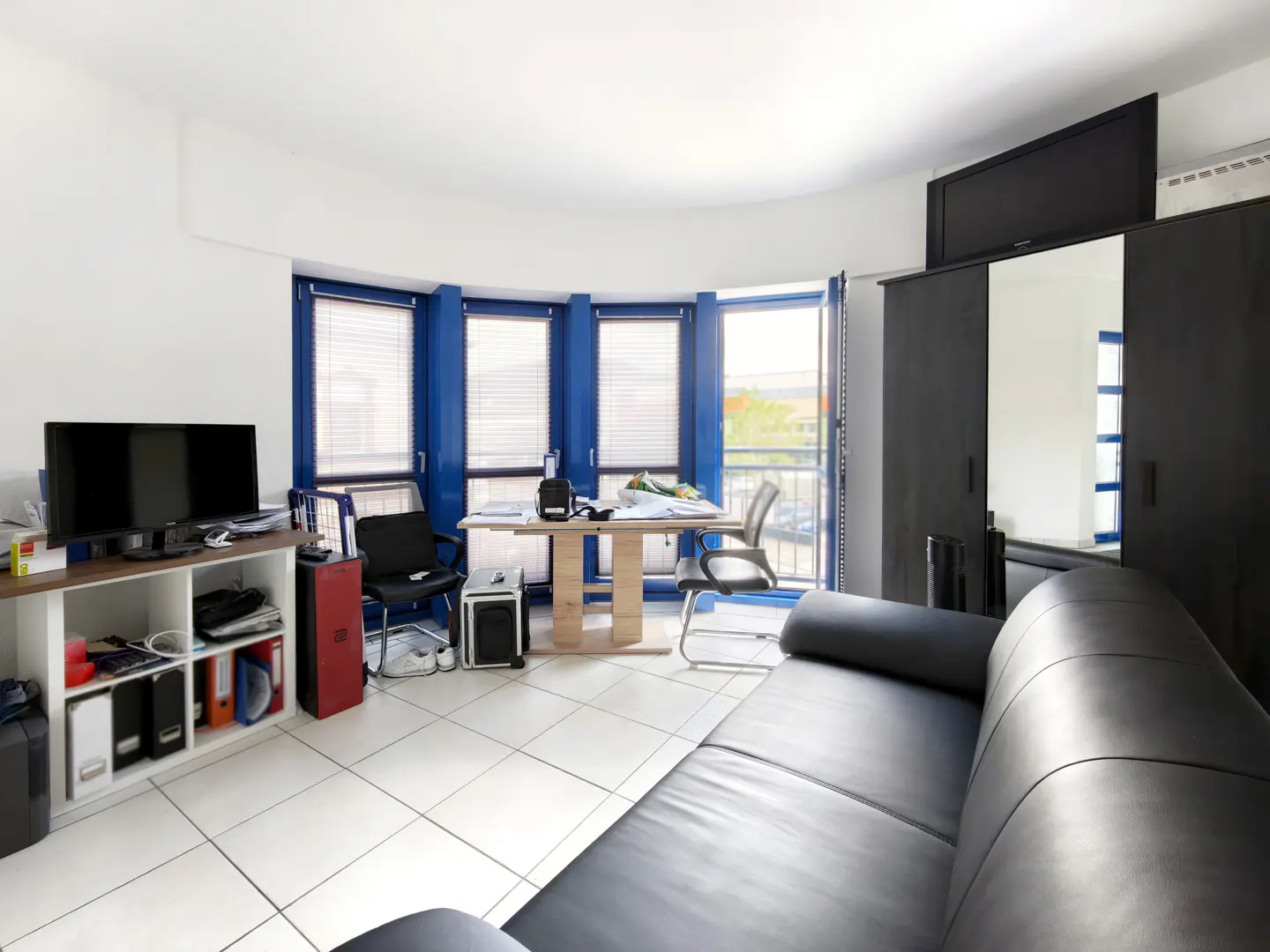 Bright home office with a black leather sofa, desk, chairs, white shelving unit, and blue-framed windows with blinds.