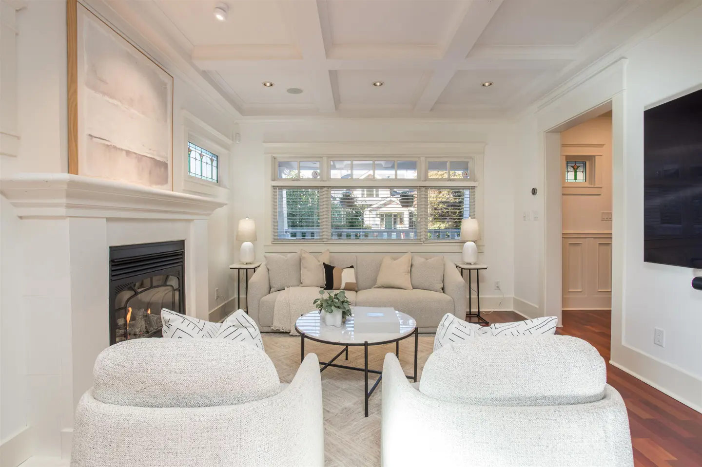Bright living room with white walls, coffered ceiling, and fireplace. Two white chairs face a sofa and round table. Large window with blinds.