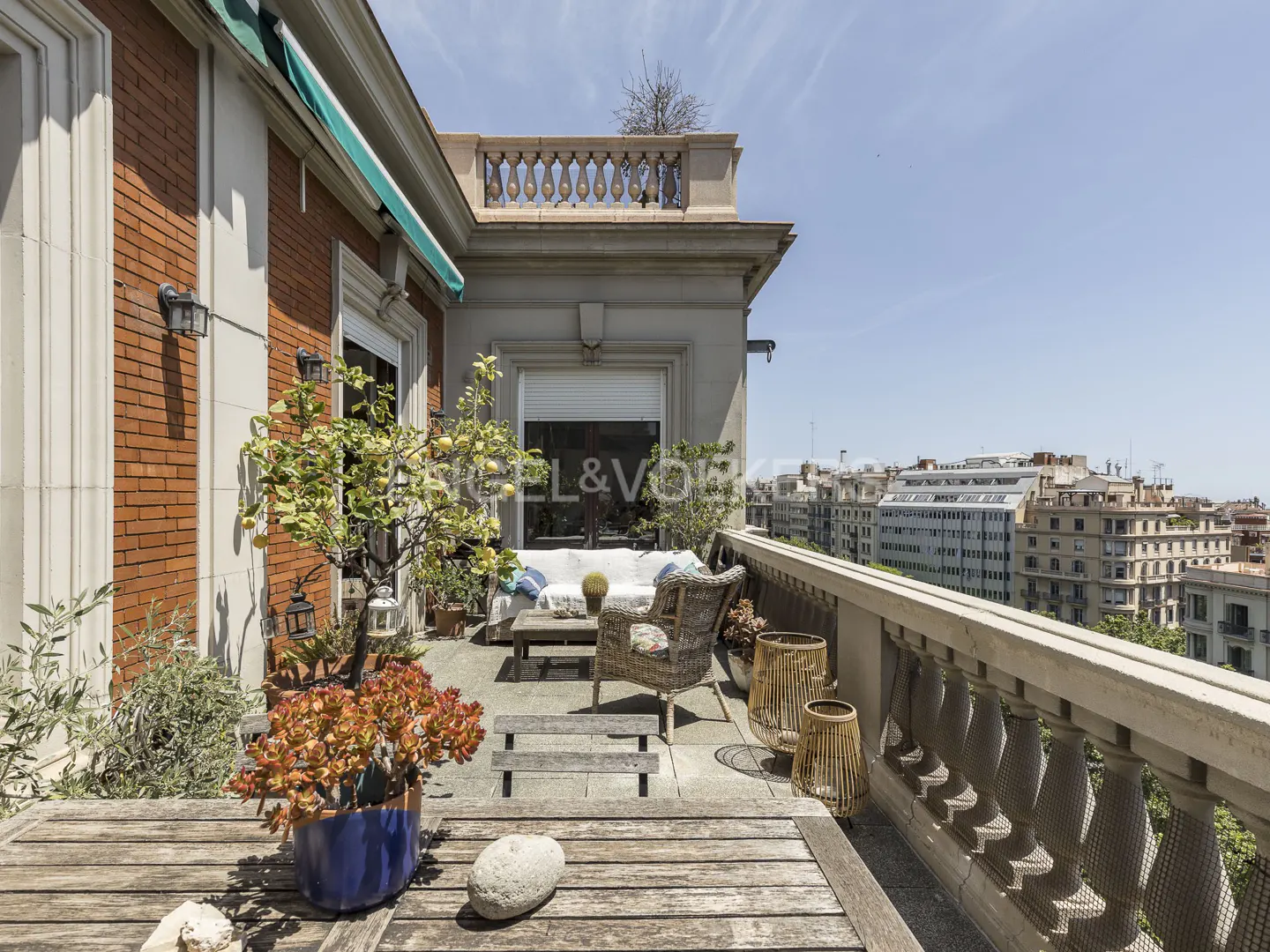 Spacious sunny terrace with wicker furniture, potted plants, and a stone balustrade overlooking a city skyline.