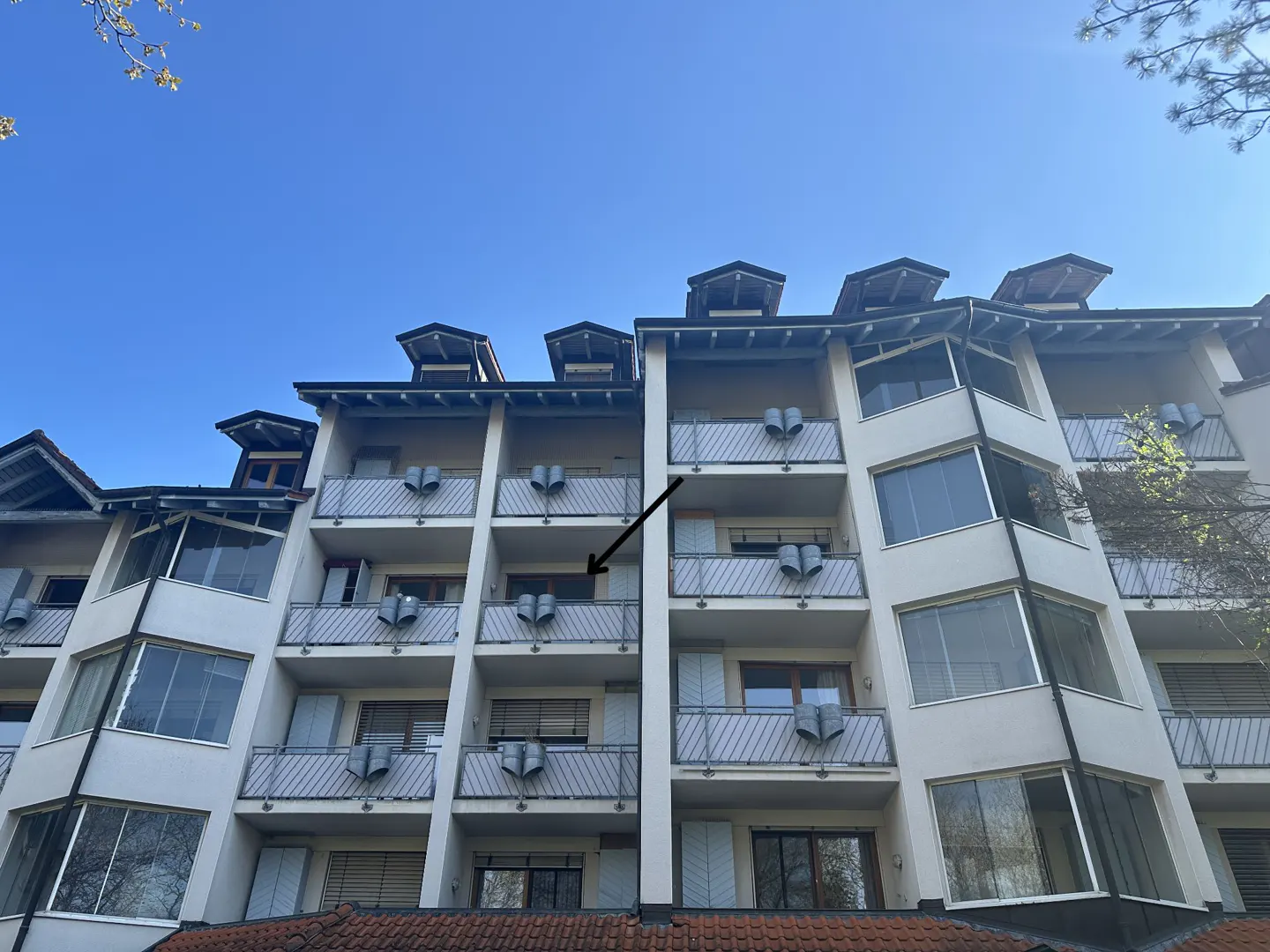 Apartment building exterior with balconies and white shutters under a blue sky. An arrow points to one of the balconies.