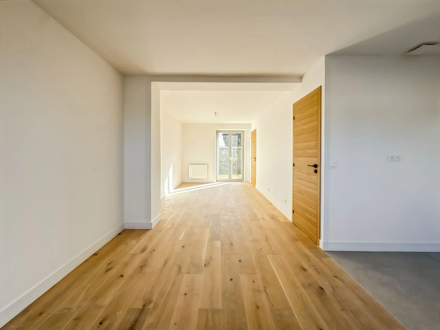Bright, empty room with light wood floors, white walls, and a wood door. Sunlight streams in from a window in the back.