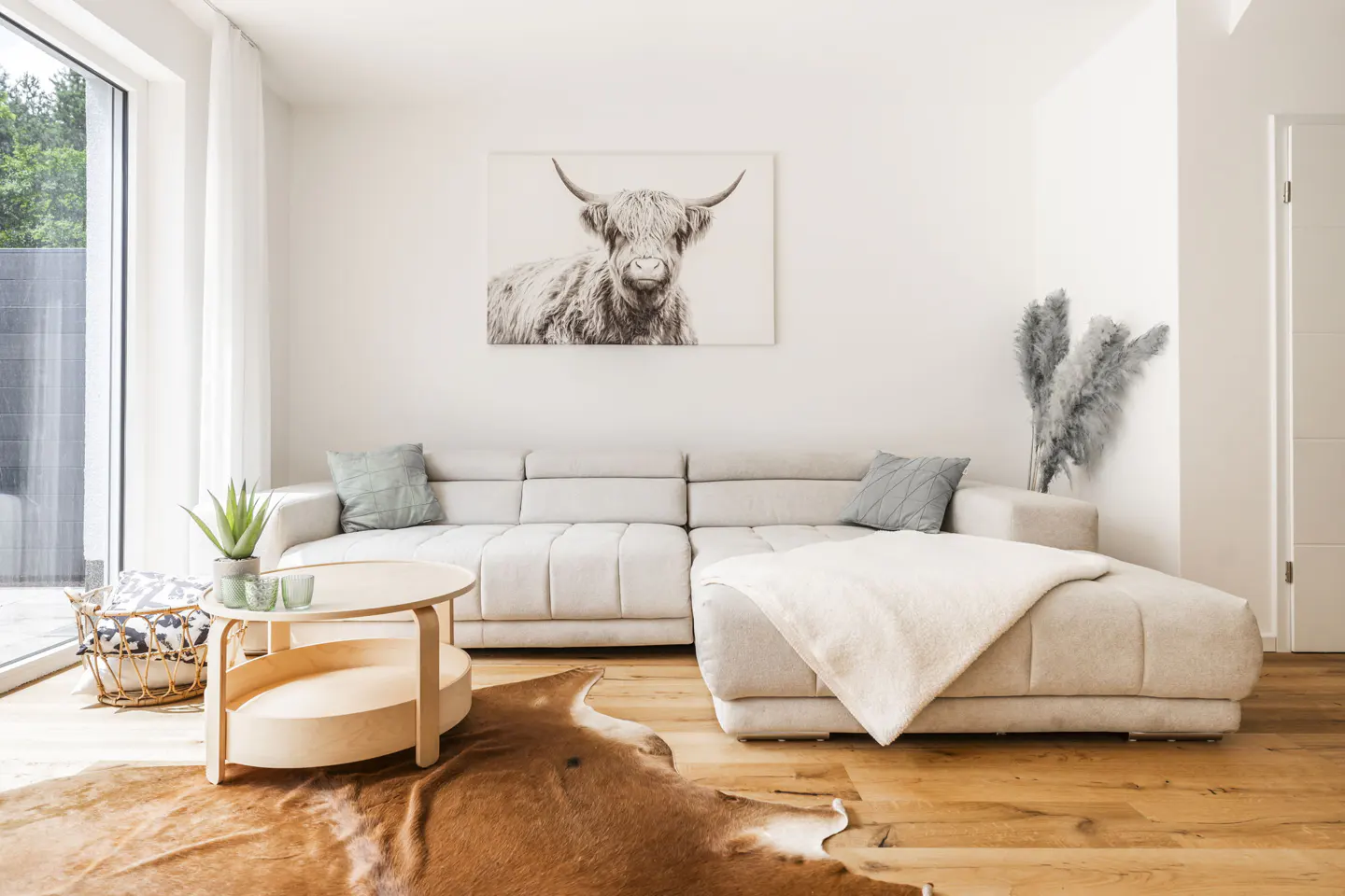 Bright living room with a beige sectional sofa, a round wooden table, and a cowhide rug on a wood floor. A Highland cow print hangs on the white wall.