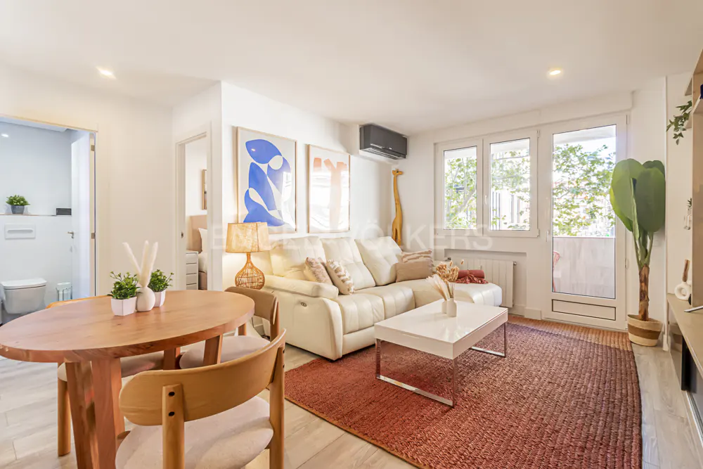 Bright living room with a round wooden table, white sofa, and a red rug. Art prints hang on the white wall.