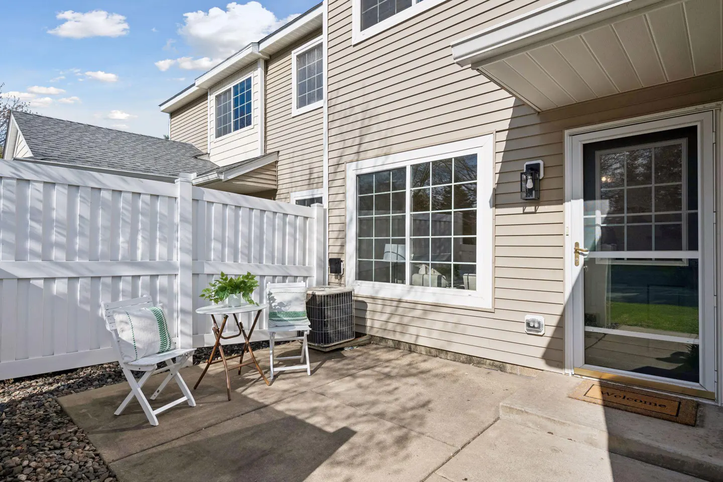 Exterior view of a beige house with a white fence, chairs, and a table on the patio. A welcome mat is in front of the door.