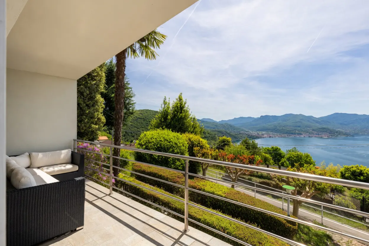 Balcony view with a black wicker sofa and white cushions overlooking a lake, mountains, and green trees under a blue sky.