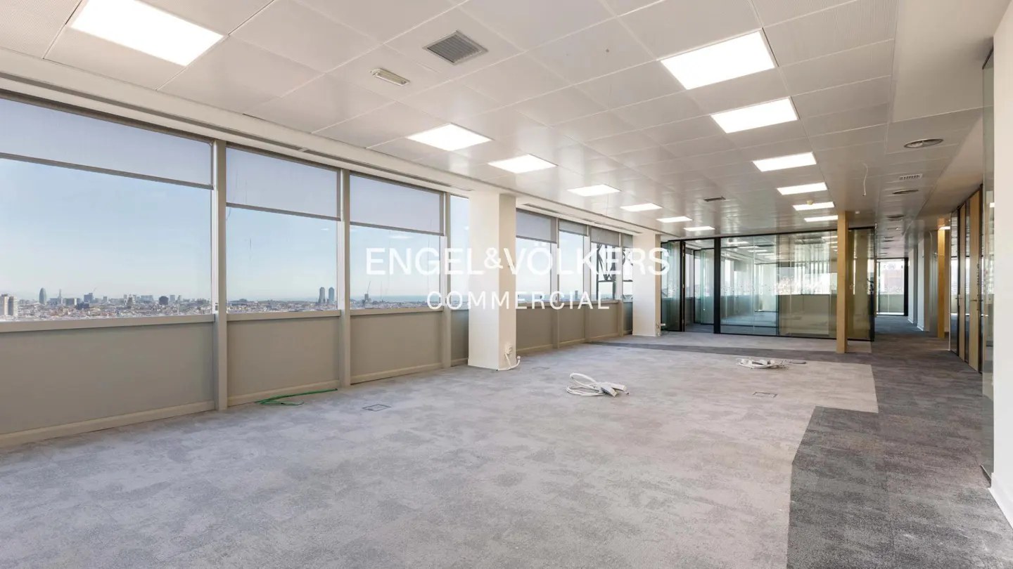 Bright, empty office space with large windows overlooking a city skyline. Gray carpet and white walls. Engel & Volkers logo on the window.