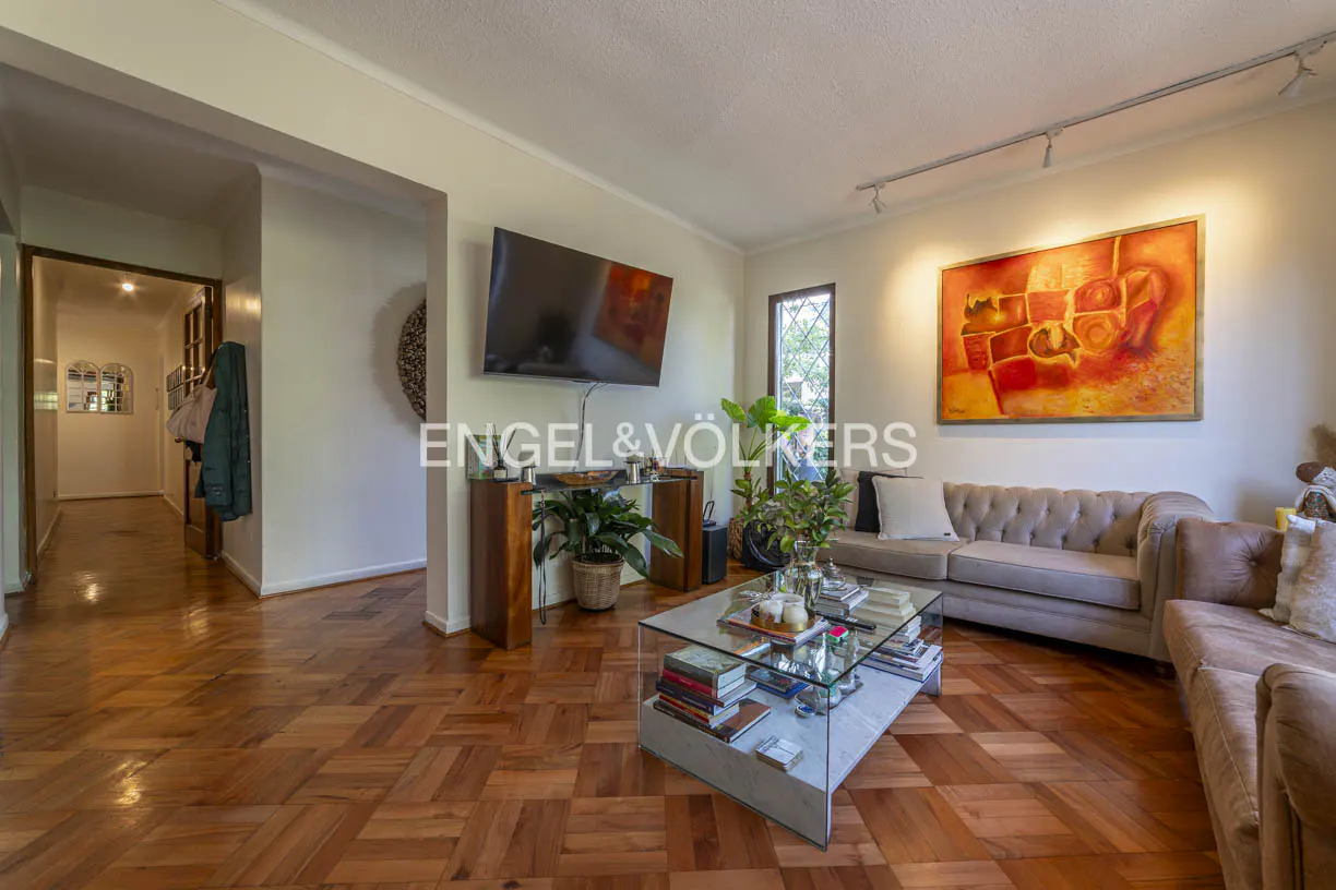 Living room with parquet floors, a glass coffee table with books, and a beige tufted sofa under an orange abstract painting.