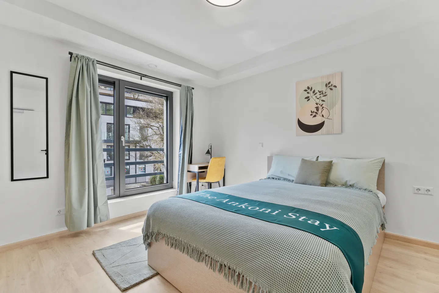 Bright bedroom with a bed, desk, and window. The bed has a light blue blanket with "The Ankoni Stay" written on it. A yellow chair sits at the desk.