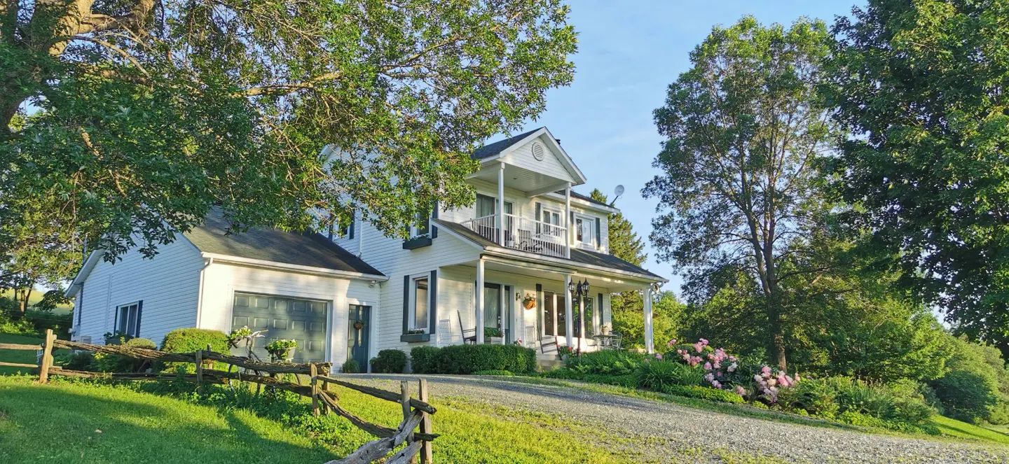 Two-story white farmhouse with a garage, porch, and balcony, surrounded by green trees and a wooden fence.