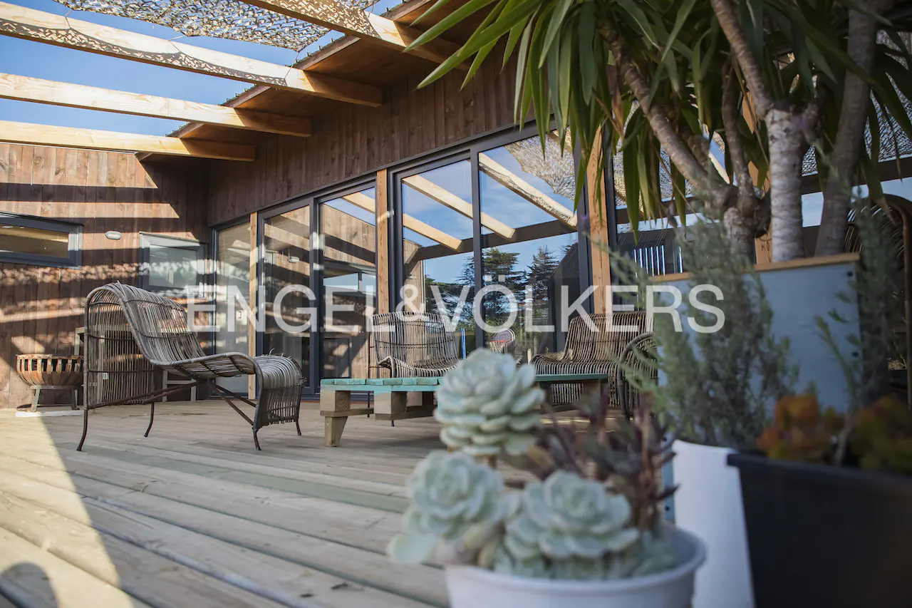 Modern wooden deck with wicker chairs, a low pallet table, and potted succulents under a slatted pergola on a sunny day.