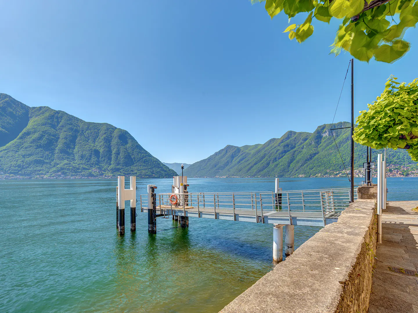 Scenic view of a lake with mountains, a pier, and a stone wall under a clear blue sky.