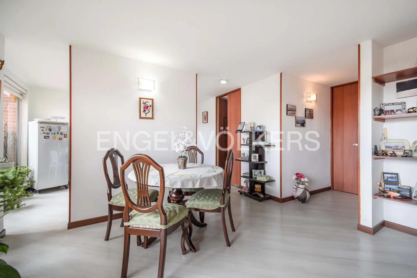 A bright dining room with a round table, four chairs, and white walls. Shelves display photos and decor. A doorway leads to the kitchen.