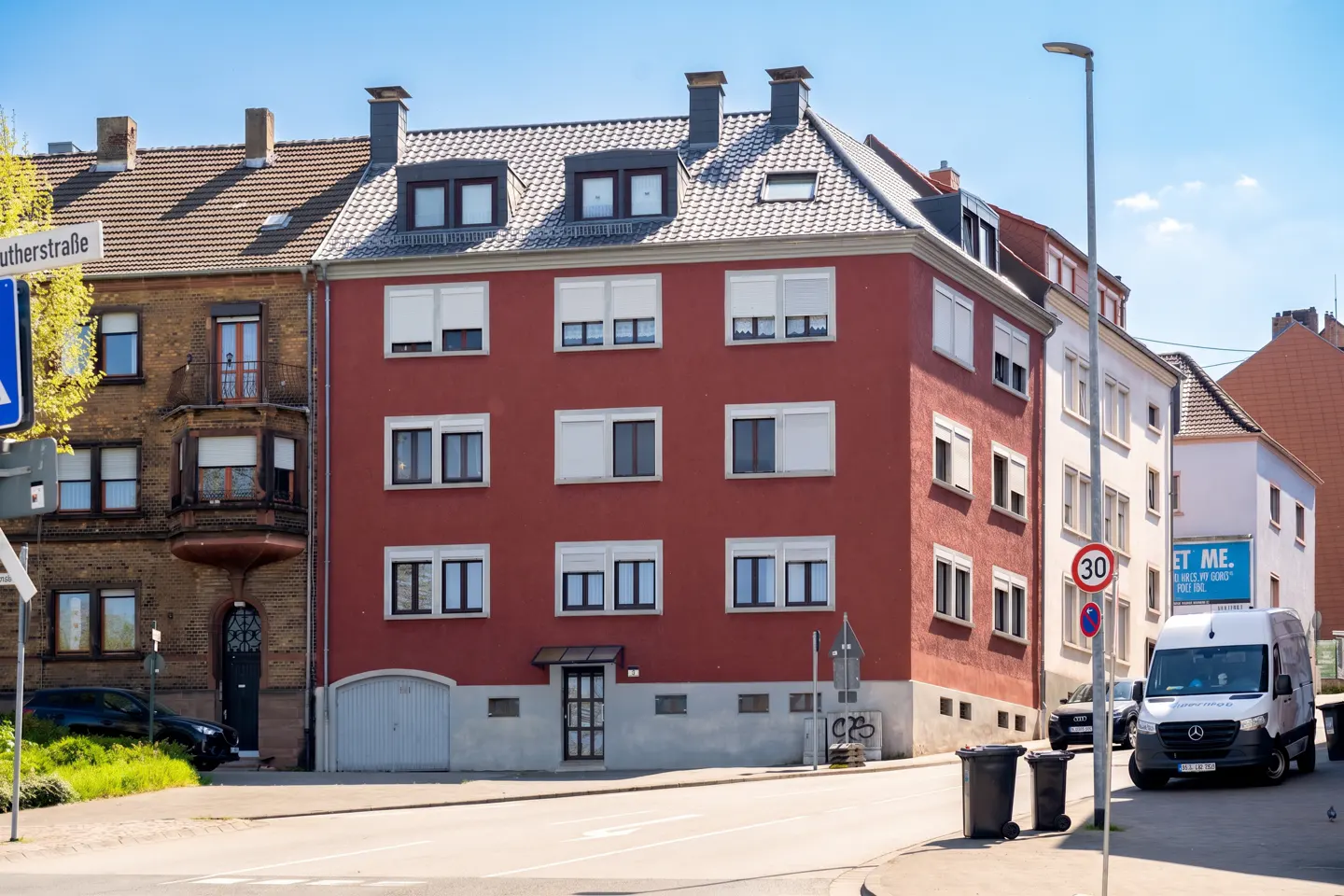 A four-story red apartment building on a street corner with white window shutters, a grey garage door, and a white van.