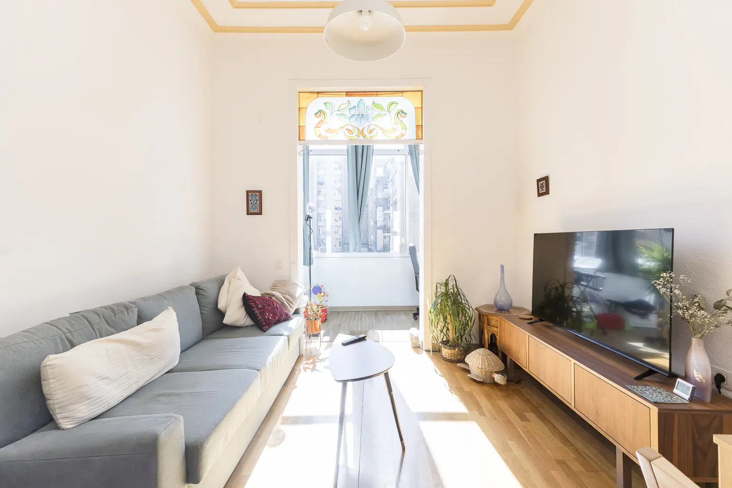 Bright living room with a gray sofa, wooden floors, and a TV on a wooden stand. A stained-glass window is above the doorway.