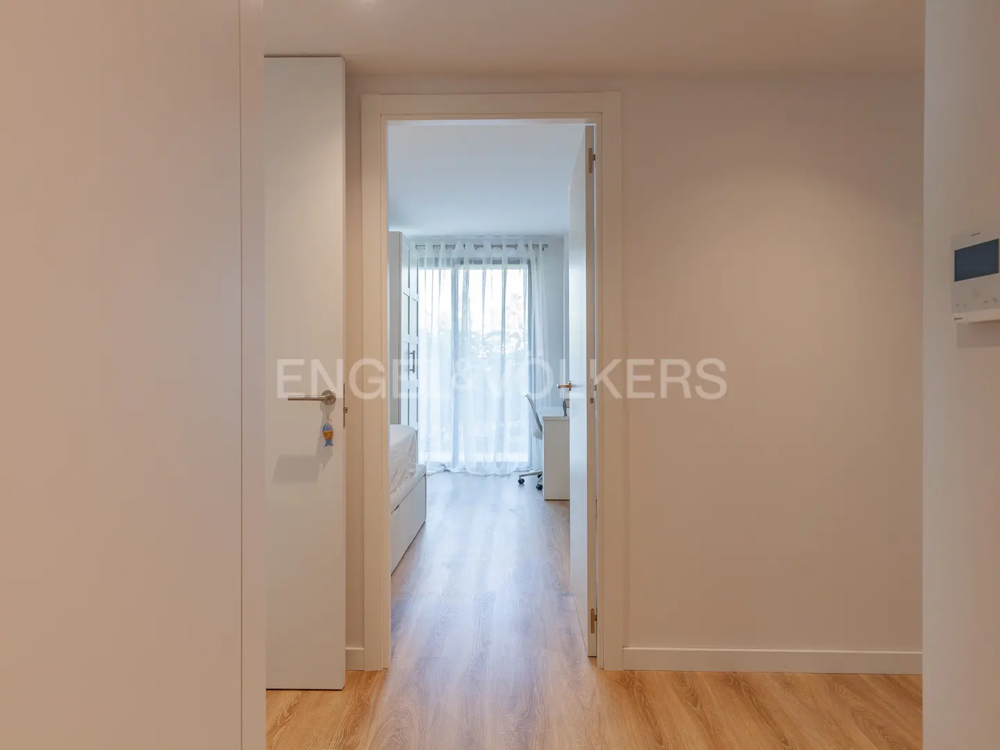A bright, modern room with wood floors is seen through an open doorway. A bed, desk, and sheer curtains are visible. The walls are white.