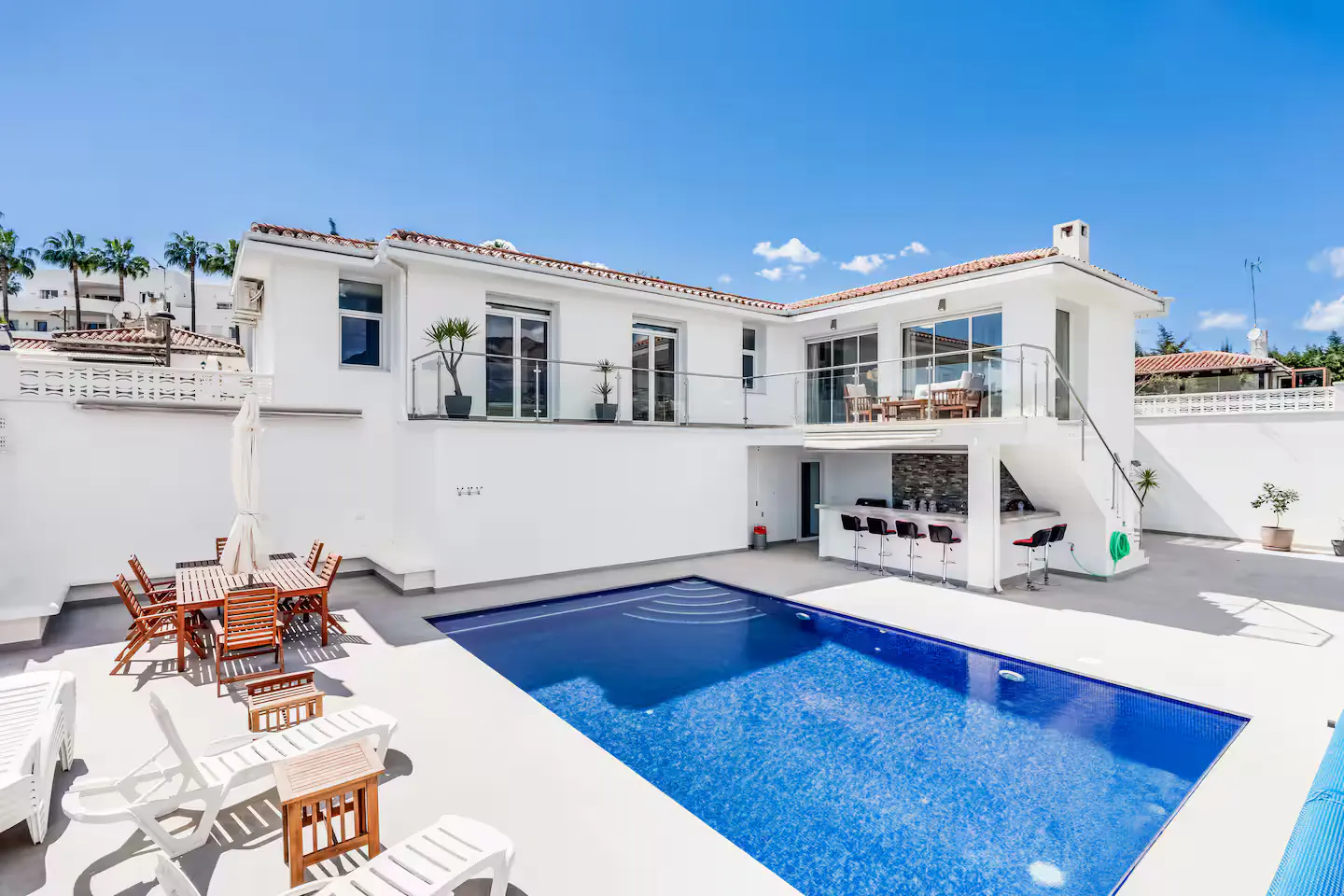 Exterior view of a modern white house with a blue swimming pool, outdoor bar, and patio furniture under a clear blue sky.