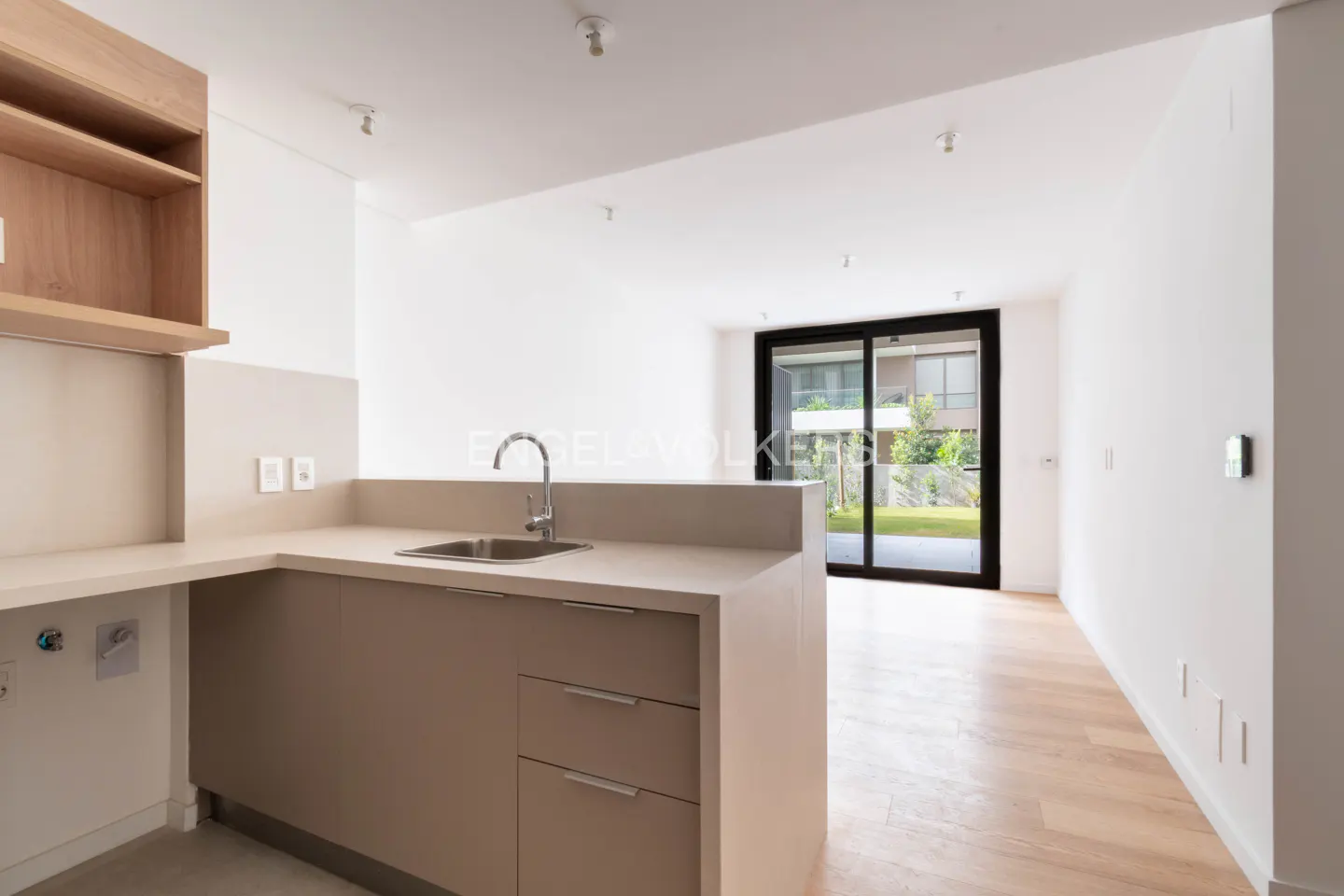 Modern kitchen with beige cabinets, stainless steel sink, and light wood floors, opening to a bright living area and patio.