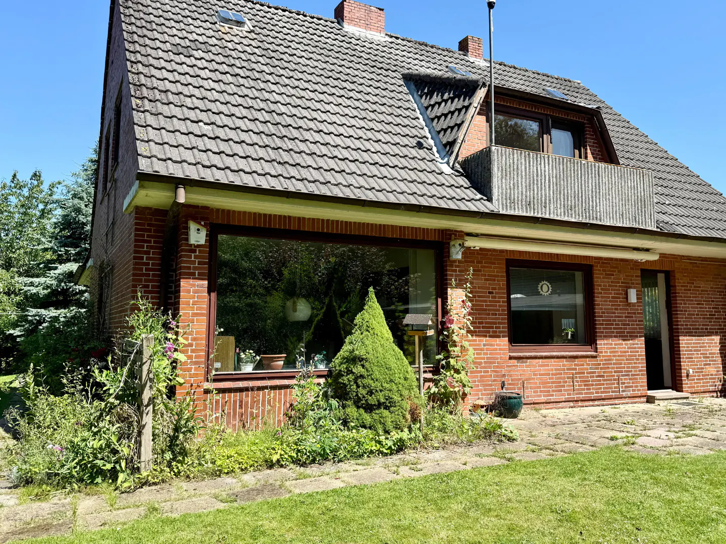 A two-story brick house with a gray tiled roof, a balcony, and a large window is surrounded by green grass and plants.
