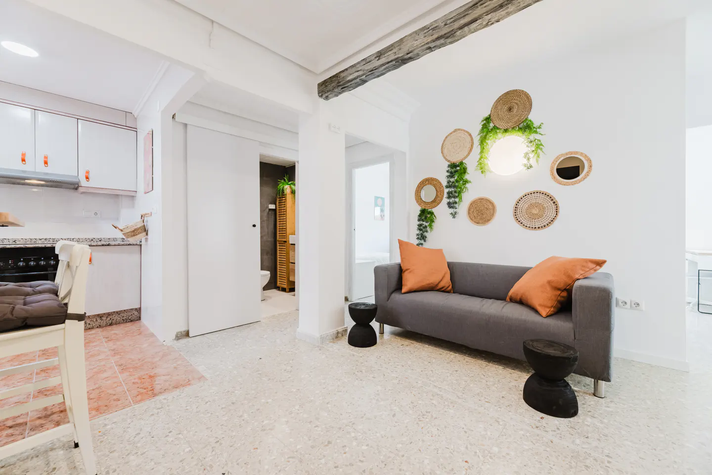 Interior view of a modern apartment with a gray sofa, orange pillows, and woven wall decor. Kitchen and bathroom visible in the background.