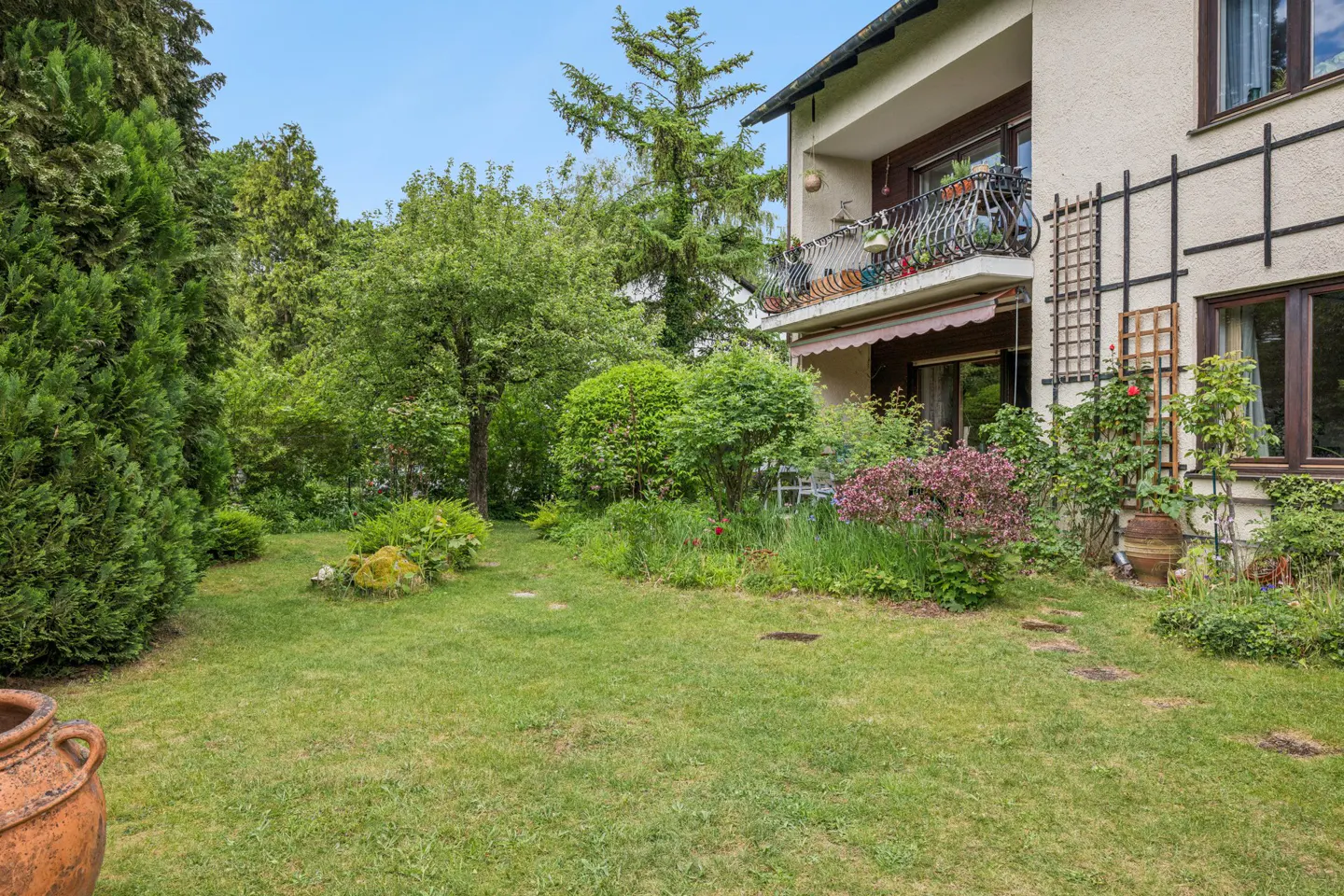 Lush green backyard with a lawn, mature trees, flowering shrubs, and a house with a balcony and decorative wall trellis.