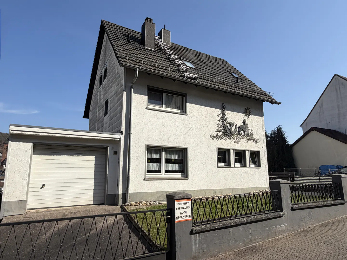 Two-story white house with a gray roof and a garage on the left. A black metal fence surrounds the property. A decorative metal art piece is on the right side of the house.