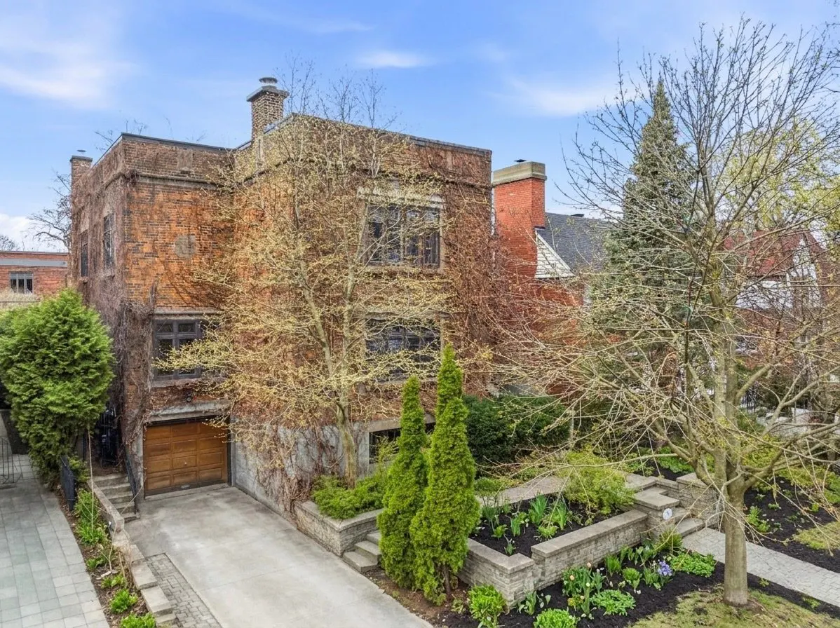 Exterior view of a large, red brick house with a wooden garage door and climbing vines. The house has a chimney and is surrounded by trees and landscaping.