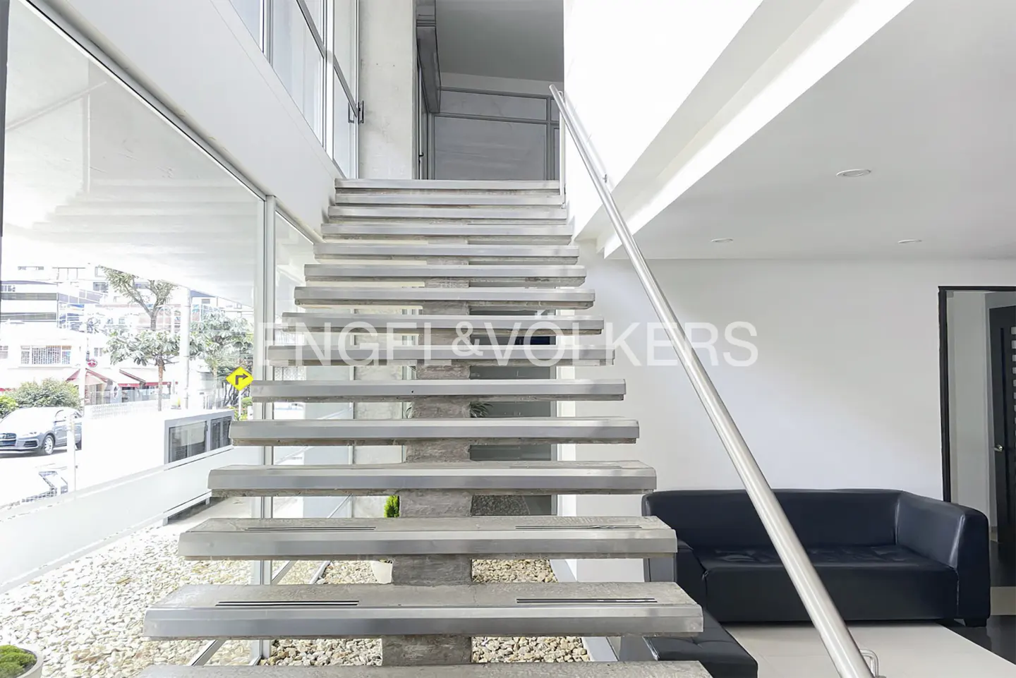 Modern interior with floating metal stairs, a stainless steel handrail, and a black leather sofa. Large windows overlook a city street.
