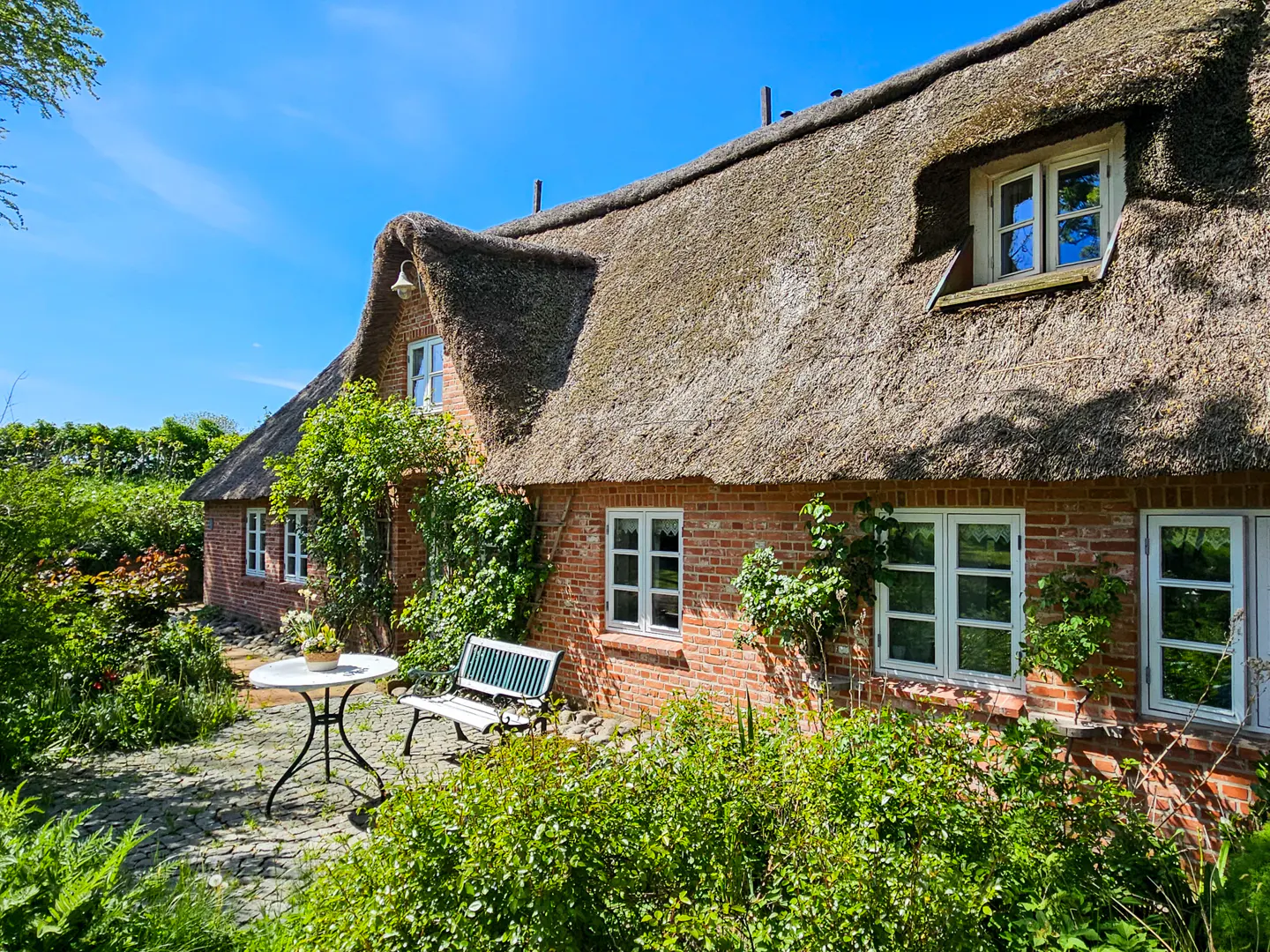 Exterior of a red brick house with a thatched roof, white windows, and a garden with a table and bench.