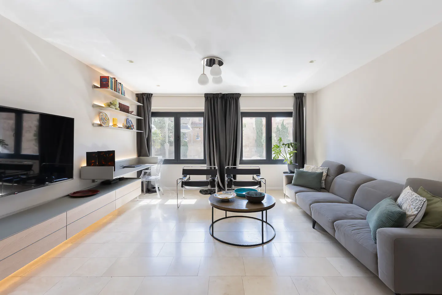 Bright living room with a gray sofa, round coffee table, and two black chairs. Large windows with dark curtains let in natural light. A TV and shelves are on the left wall.
