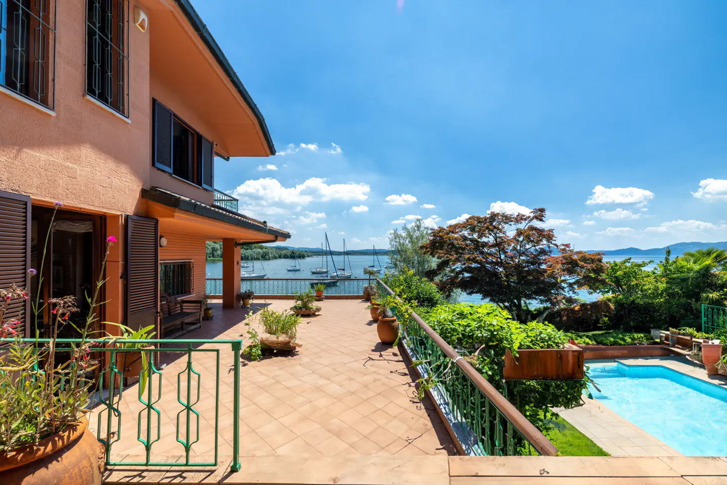 Exterior view of a peach-colored house with a tiled patio, green railings, and a pool overlooking a lake with sailboats under a blue sky.