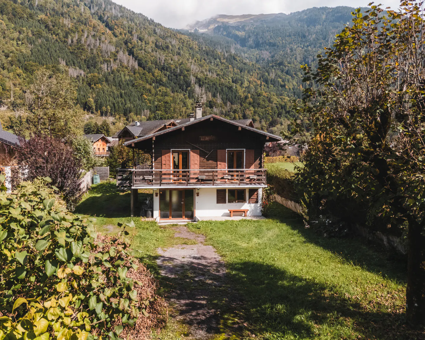 Two-story chalet with a brown wood upper level and white lower level, surrounded by green grass and trees. Mountains in the background.