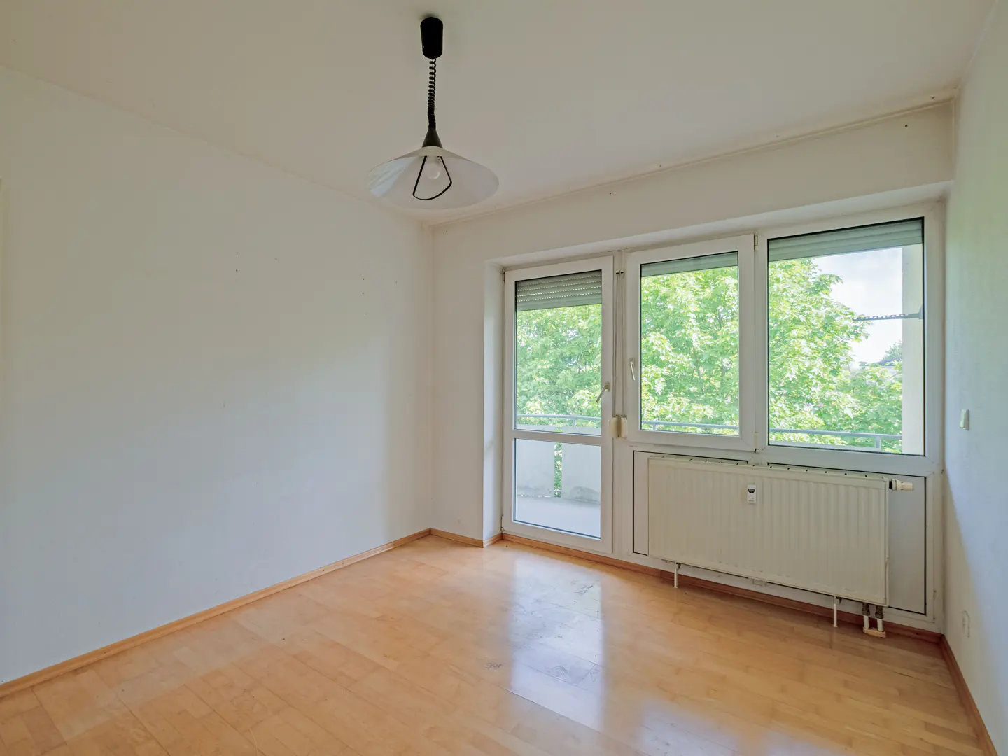 Empty room with light wood floors, white walls, a pendant light, and a large window overlooking green trees.