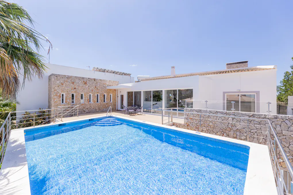 A bright blue pool sits in front of a modern white house with stone accents and large windows. Palm tree on the left.