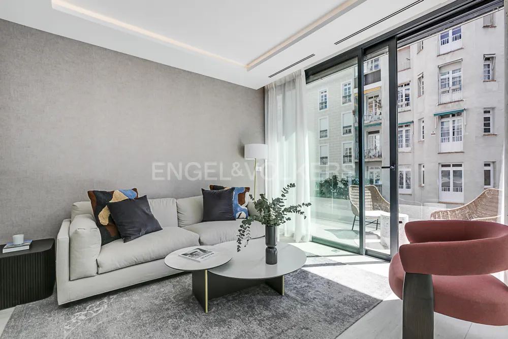 Living room with a white sofa, pillows, and a round coffee table. Sliding glass doors open to a balcony with chairs and a view of a building.