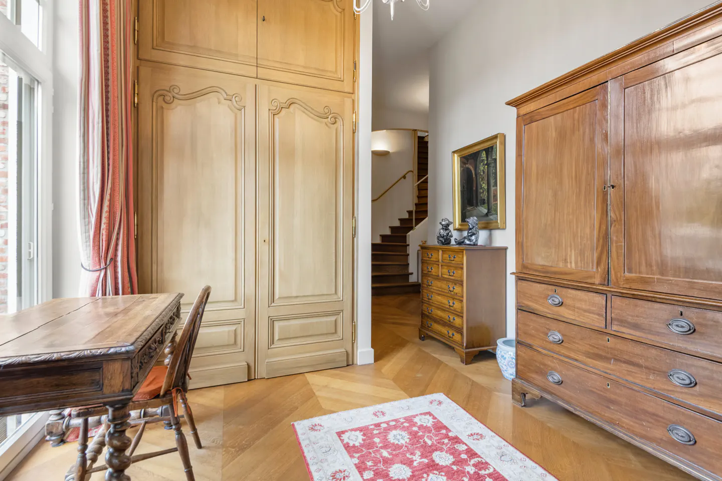 A room with wooden floors, a desk, a chair, and a large wooden wardrobe. A staircase is visible in the background.