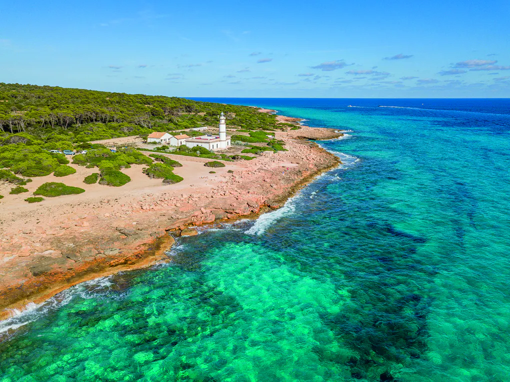 Aerial view of a white lighthouse on a rocky coast with turquoise water and a blue sky.