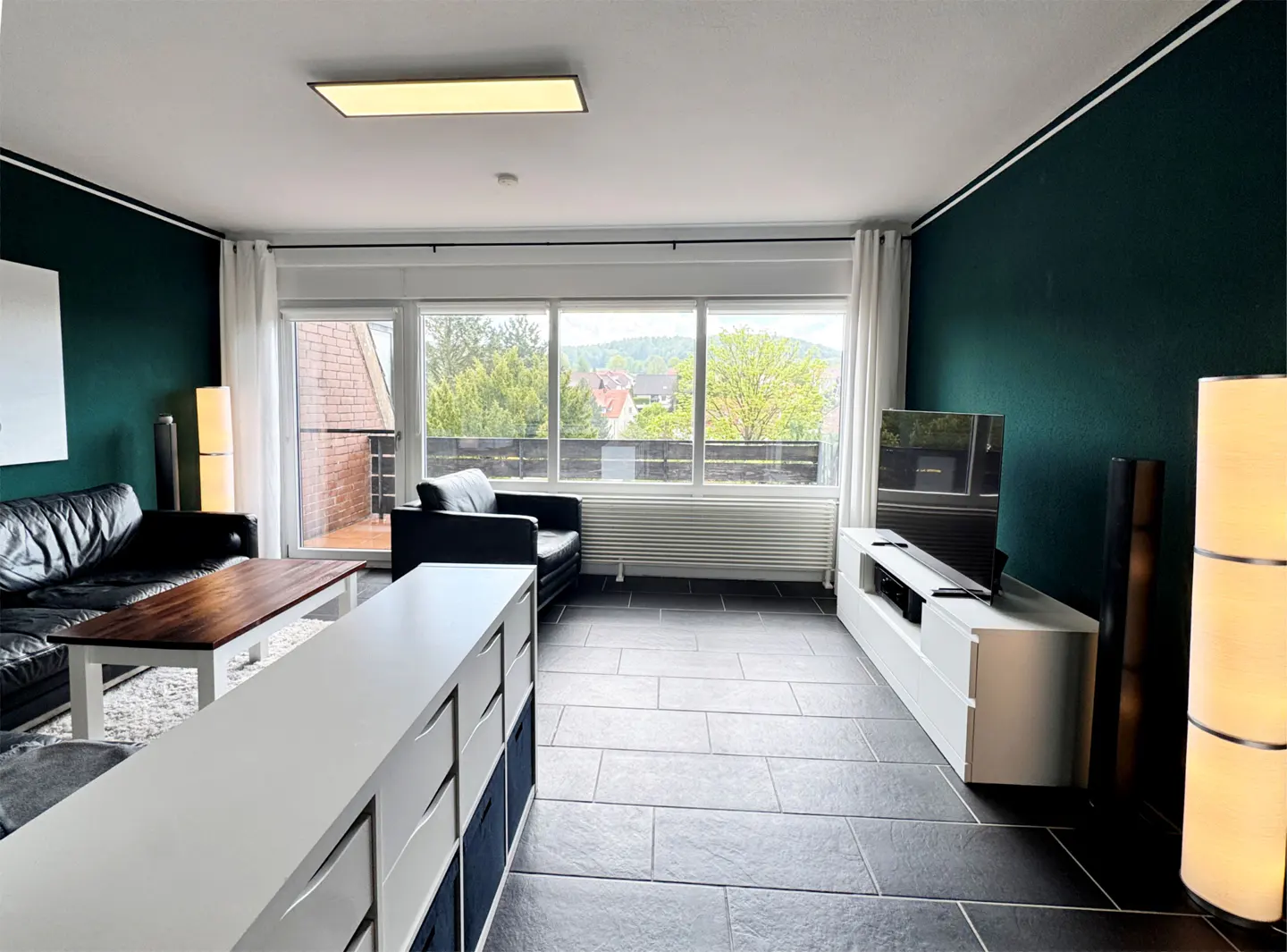 A living room with dark green walls, black tile floors, and large windows. Furnishings include a black leather sofa and chair, a white storage unit, and a TV.