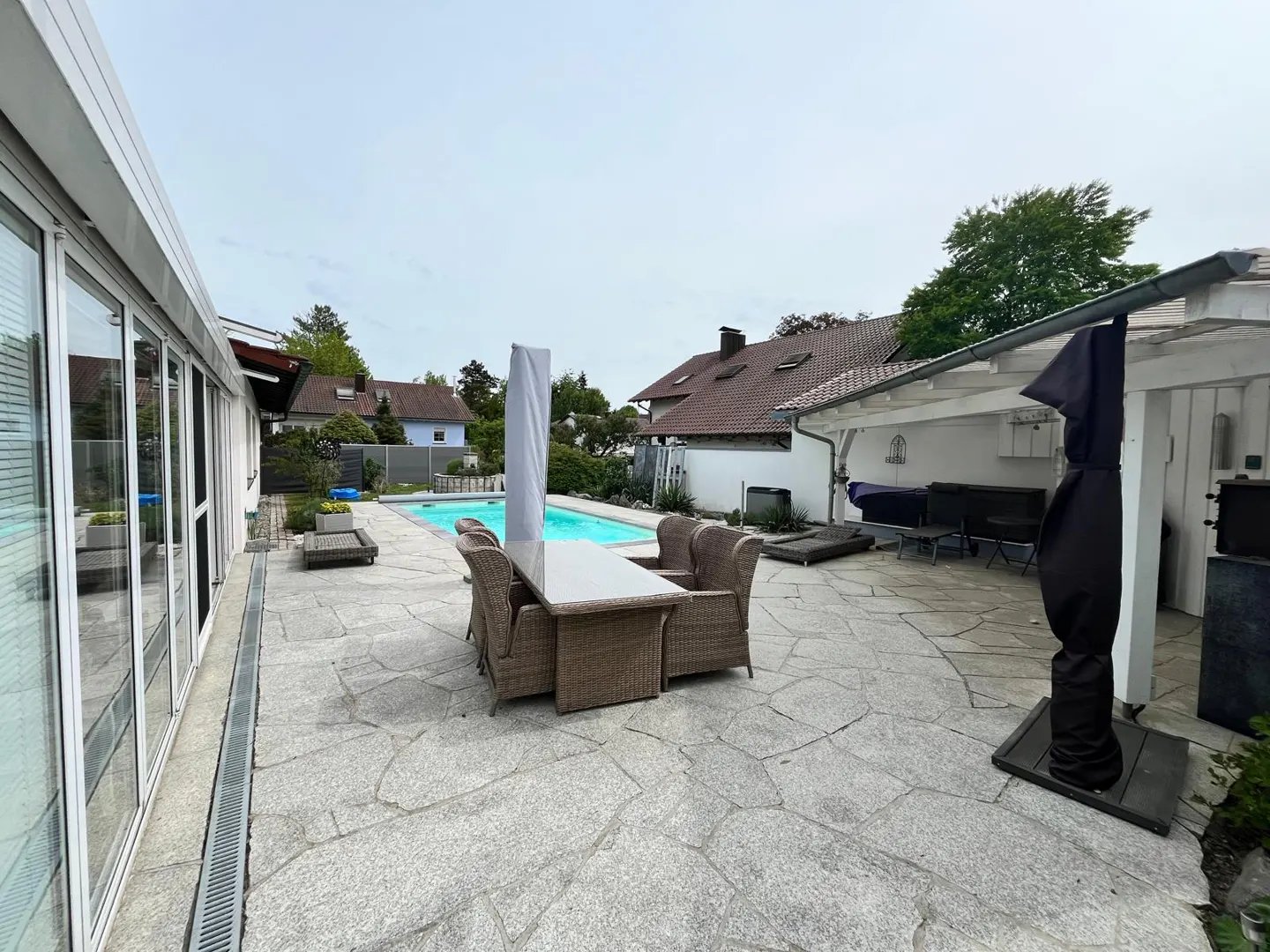 Outdoor patio with a pool, table, and chairs. The patio is made of stone and there is a covered area with furniture.