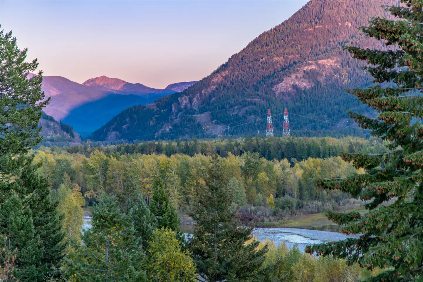 Scenic view of a valley with a river, trees, and mountains in the background at dusk. Two red and white transmission towers are visible.
